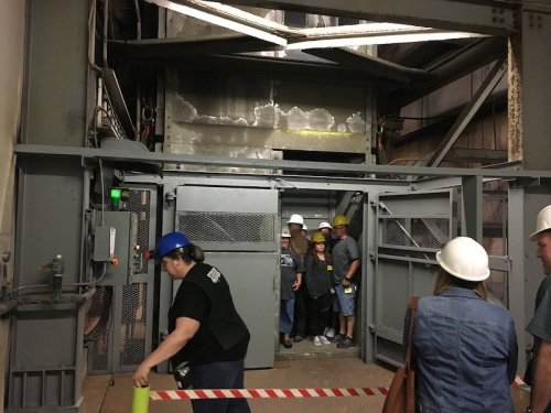 A group of people in hard hats stands near an industrial elevator in a warehouse setting.