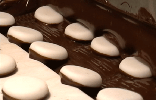 Chocolate-covered cookies on a conveyor belt, partially submerged in melted chocolate.