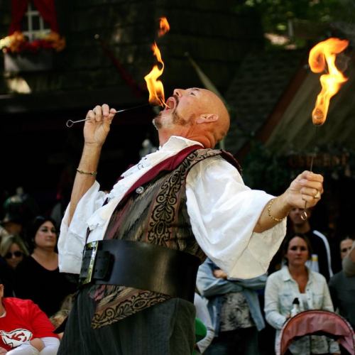A performer in a costume breathes fire, captivating an audience at an outdoor festival.