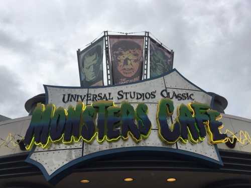 Sign for Universal Studios' Classic Monsters Cafe, featuring colorful monster-themed graphics against a cloudy sky.