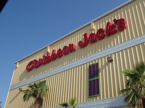 Sign for Caribbean Jack's restaurant on a yellow building with palm trees in front, under a clear blue sky.
