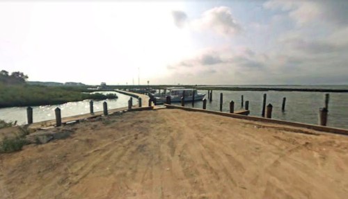 A sandy area leading to a dock with boats, surrounded by water and cloudy skies.