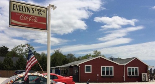 A red building with a sign reading "Evelyn's Drive In," surrounded by cars and a flag, under a blue sky with clouds.