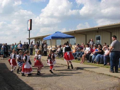 A group of girls in traditional costumes dance in front of an audience seated outside a building under a blue sky.