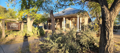 A cozy brick house with a porch, surrounded by trees and plants, featuring a sign for a pizza restaurant.