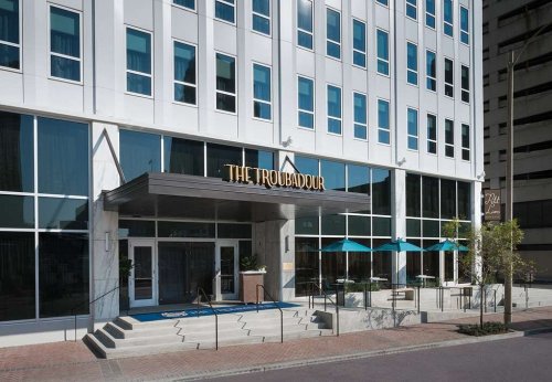 Modern hotel entrance with large glass windows, a sign reading "The Troubadour," and blue awnings over the entrance.