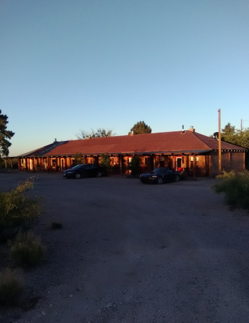 A rustic building with a red roof and multiple entrances, surrounded by gravel and sparse vegetation under a clear sky.