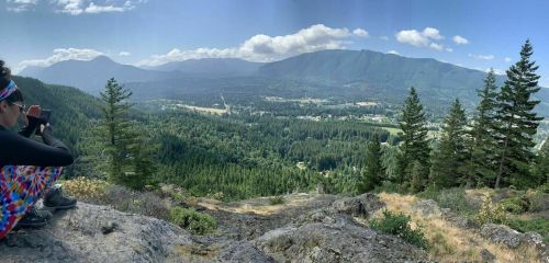 A person in colorful pants sits on a rocky ledge, taking photos of a lush green valley and mountains in the distance.