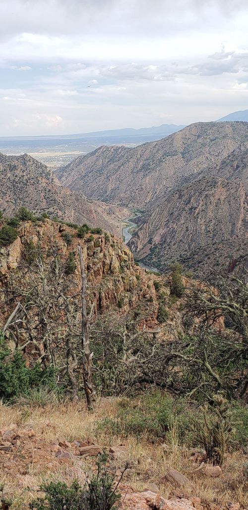 A scenic view of a winding river through rugged mountains, with dry vegetation in the foreground and cloudy skies above.