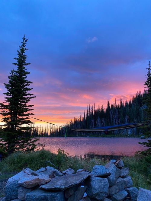A serene lakeside scene at sunset, featuring a hammock between trees and a stone fire pit in the foreground.