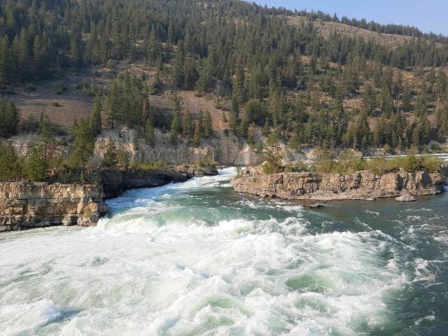 A rushing river flows through rocky terrain, surrounded by lush green trees and steep cliffs in a natural landscape.