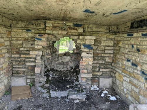 Interior of a stone structure with a large hole in the wall, surrounded by debris and graffiti on the walls.