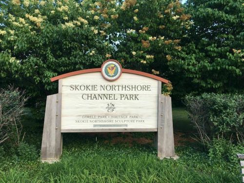 Sign for Skokie Northshore Channel Park, surrounded by greenery, featuring park names and a logo.