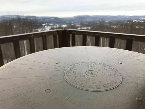 A metal survey marker on a wooden railing, overlooking a scenic winter landscape with distant hills and valleys.