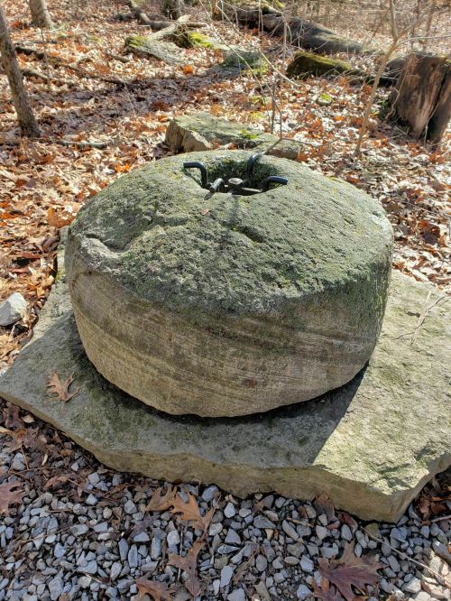 A large, moss-covered stone millstone sits on a rocky surface in a wooded area.