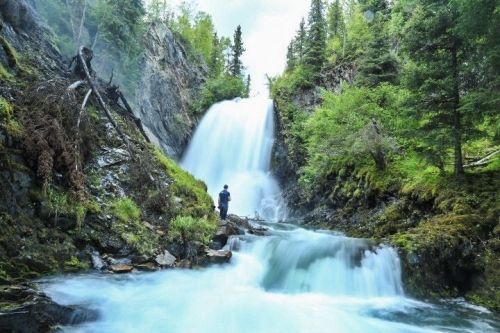 A person stands near a cascading waterfall surrounded by lush greenery and rocky terrain.