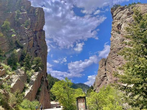 A scenic view of a narrow canyon with towering rock formations, lush greenery, and a bright blue sky with clouds.