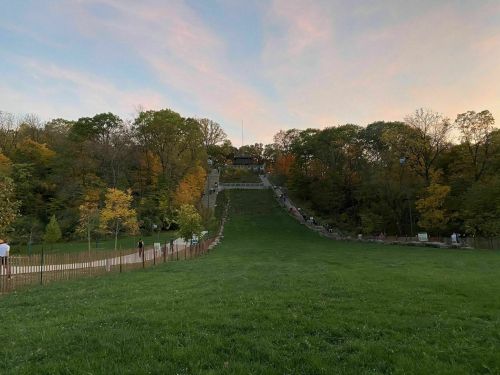 A grassy hill leads to a staircase surrounded by trees, with people walking up as the sun sets in the background.