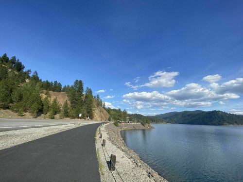 A scenic view of a calm lake beside a paved road, surrounded by green trees and mountains under a blue sky with clouds.
