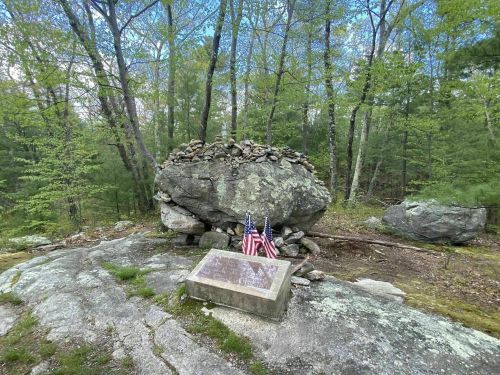 A large rock with a stone pile on top, featuring a plaque and two American flags, surrounded by trees.