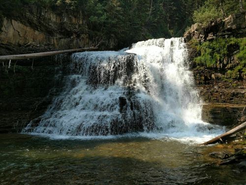 A cascading waterfall flows over rocky tiers into a serene pool, surrounded by lush greenery and forested cliffs.