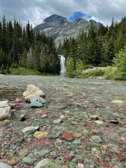 A clear river with colorful stones flows toward a waterfall, surrounded by lush trees and mountains under a cloudy sky.