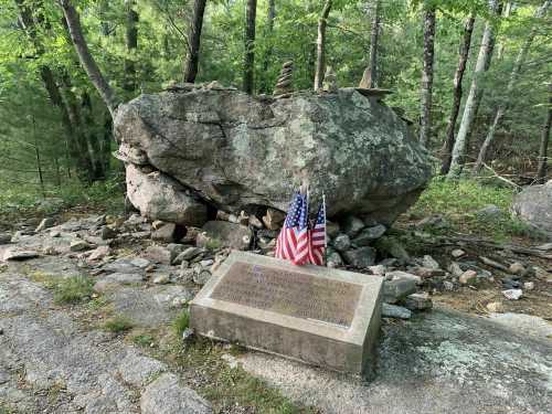 A large rock with a plaque and two American flags, surrounded by trees and stones in a natural setting.