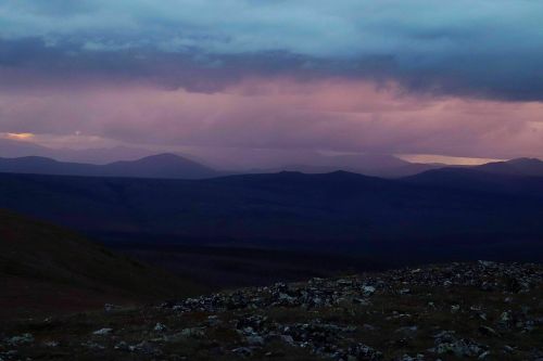 A dramatic landscape at dusk, featuring layered mountains under a cloudy sky with hints of purple and orange hues.