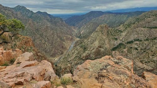 A panoramic view of a rocky canyon with a winding river below, surrounded by mountains under a cloudy sky.