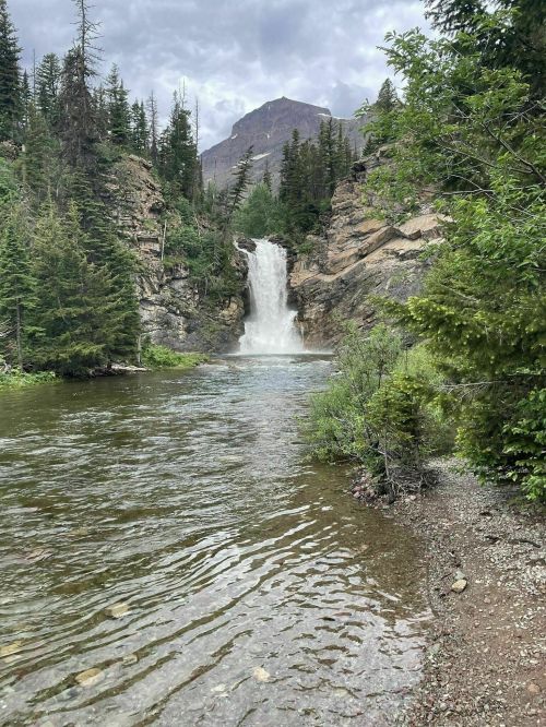 A serene waterfall cascades into a clear river, surrounded by lush greenery and rocky cliffs under a cloudy sky.