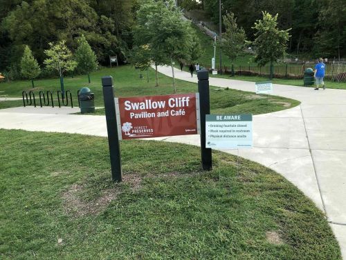 Sign for Swallow Cliff Pavilion and Café, with park path and greenery in the background. Safety notices visible.