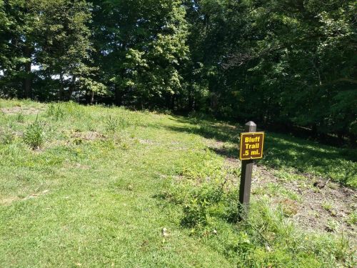 A grassy path leads into a wooded area, marked by a sign indicating the Bluff Trail is 0.5 miles ahead.