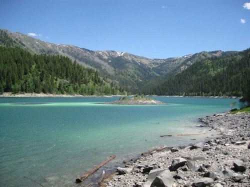 A serene lake surrounded by mountains and lush greenery, with clear blue water and a rocky shoreline.