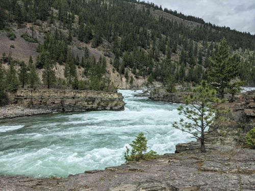 A rushing river flows between rocky cliffs, surrounded by lush green trees and mountains under a cloudy sky.