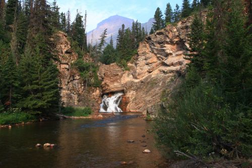 A serene waterfall cascades into a calm river, surrounded by tall trees and rocky cliffs under a clear blue sky.
