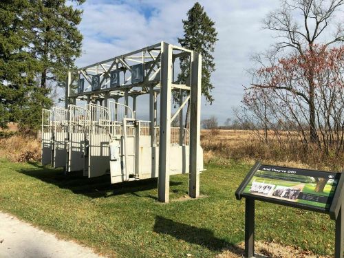 Starting gate for horse racing, with a sign nearby, set in a grassy area surrounded by trees and open fields.