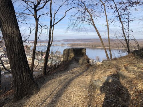 A scenic view from a rocky path overlooking a river, framed by trees and a clear sky.