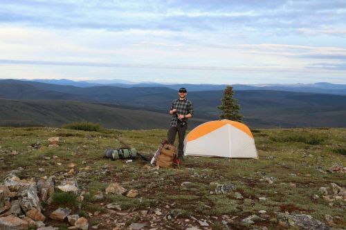 A person stands near a tent on a rocky hillside, surrounded by mountains and greenery under a cloudy sky.