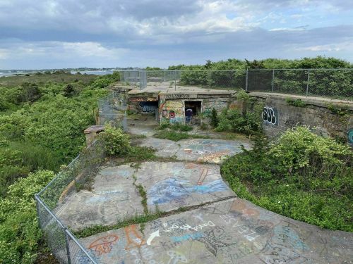 Abandoned concrete structure surrounded by greenery, featuring colorful graffiti and a chain-link fence.