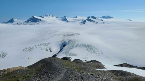 A vast, snowy landscape with distant mountains under a clear blue sky, featuring a glacier and rocky foreground.