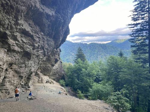 A scenic view from a cave entrance, with hikers and lush green mountains in the background under a cloudy sky.
