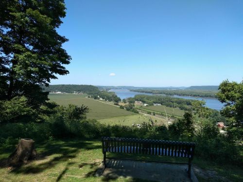 Scenic view of a river valley from a hilltop, with a bench in the foreground and clear blue skies above.