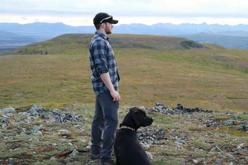 A person in a plaid shirt stands on a hillside with a black dog, overlooking a vast landscape of mountains and fields.