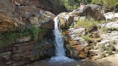 A small waterfall cascading over rocky terrain, surrounded by greenery and sunlight.