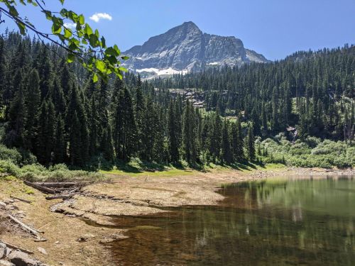 A serene mountain landscape with a clear lake, surrounded by lush green trees and a towering peak in the background.