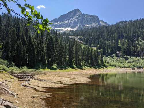 A serene mountain landscape with a clear lake, surrounded by lush green trees and a towering peak in the background.