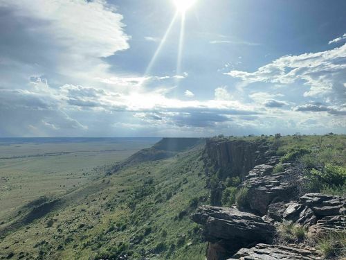 A sunlit landscape with rocky cliffs, green hills, and a vast sky filled with clouds.