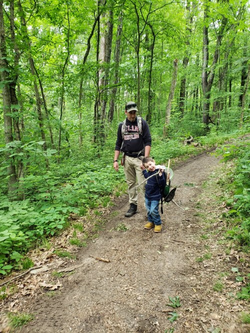 A man and a child walk along a wooded trail, surrounded by lush green trees and foliage.