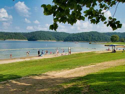 A scenic beach with people enjoying the water, surrounded by green hills and a clear blue sky.