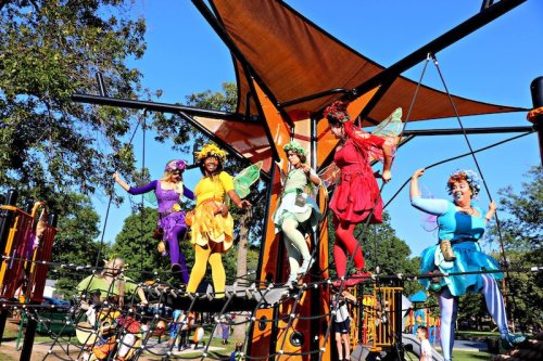 Colorful performers dressed as fairies play on a playground structure, surrounded by trees and a clear blue sky.